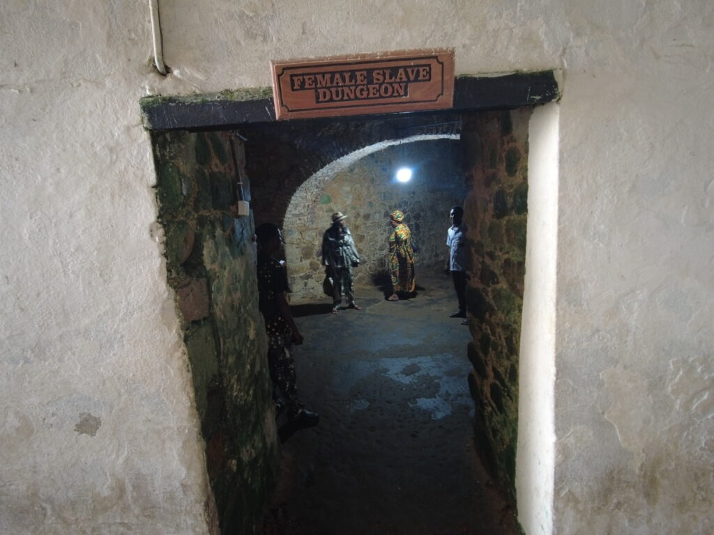 A weathered wooden sign reading "FEMALE SLAVE DUNGEON" in bold capital letters hangs above a low, narrow, arched doorway.