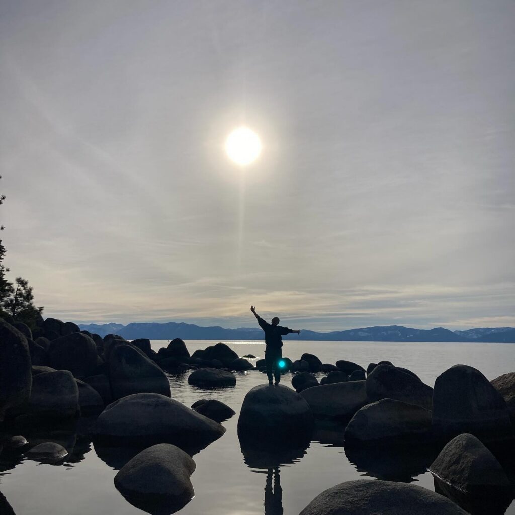 Silhouette of a person with arms outstretched on a boulder at a lake shore, sun shining over the mountains