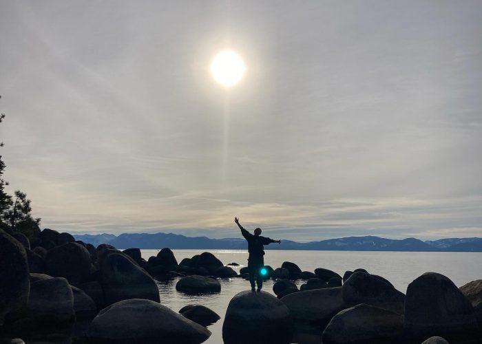Silhouette of a person with arms outstretched on a boulder at a lake shore, sun shining over the mountains