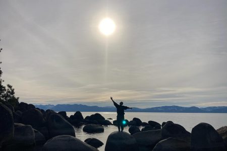 Silhouette of a person with arms outstretched on a boulder at a lake shore, sun shining over the mountains